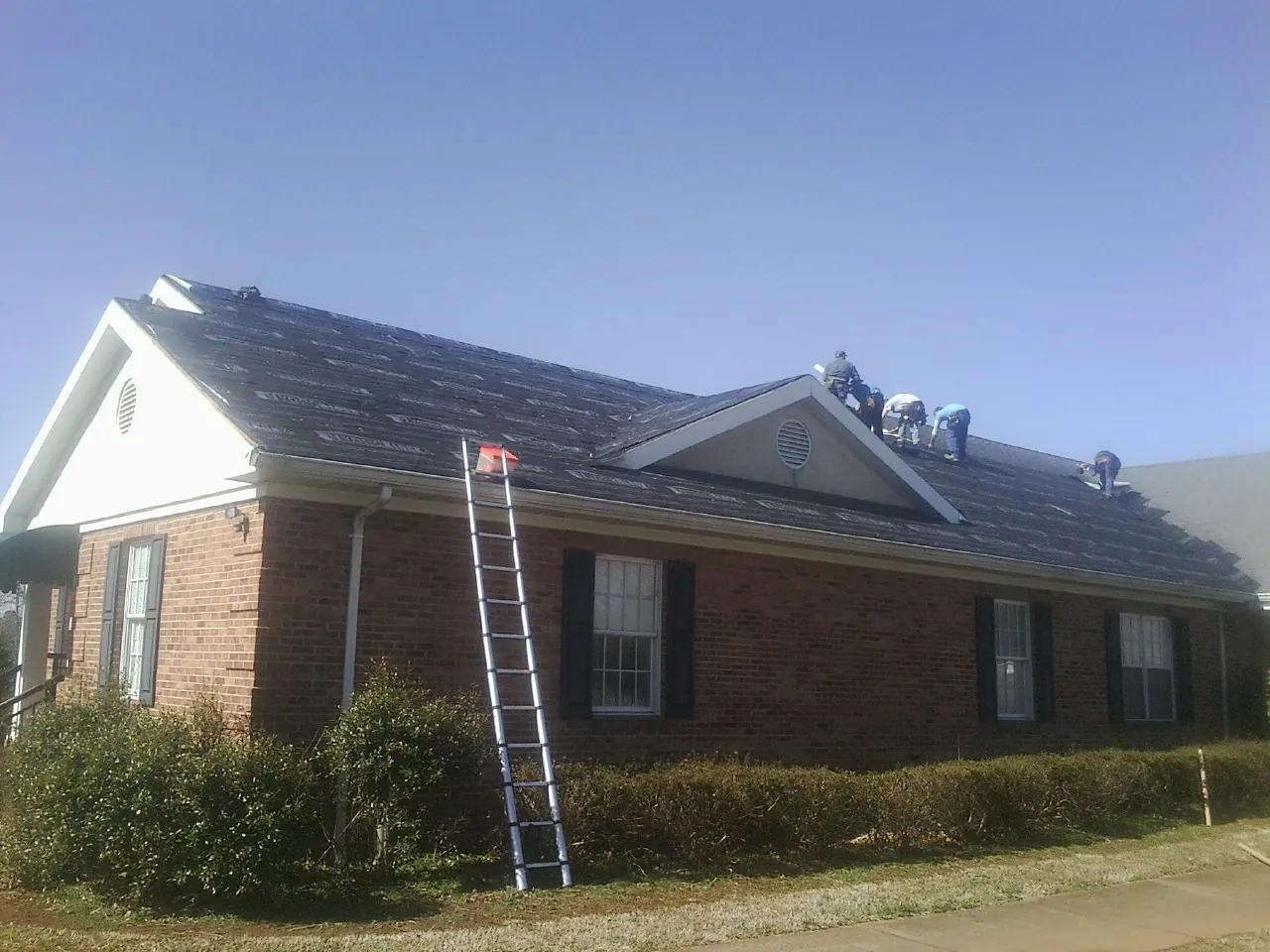 Crew tearing shingles off a brick ranch roof with a ladder set — Emergency Roof Repair work by our crew in Nemaha