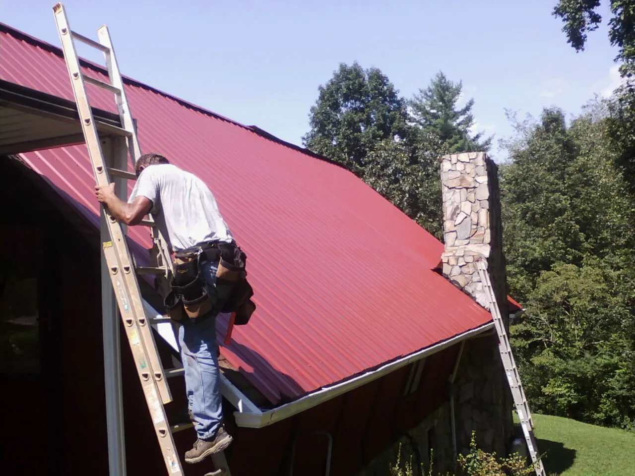 Roofer on a ladder beside a red metal roof and stone chimney — Soffit & Fascia Repair by Roofing Brothers in Nemaha